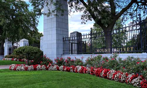 The photo shows the entrance to Mt. Olivet Cemetery, surrounded by red and white flowers.