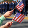 In the background are military-affiliated individuals in dark blue uniforms. In the foreground are two hands holding small American flags.