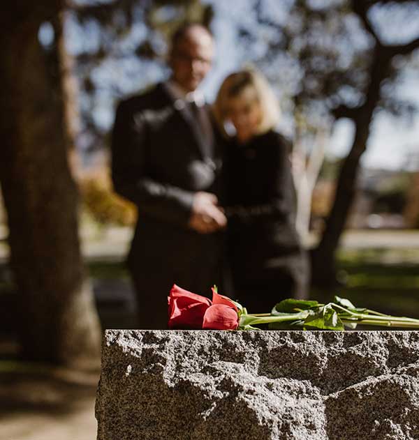 Grief expressed by a photo of a couple looking onto the grave of a loved one. A couple in the background, with a headstone in the foreground, a few roses sit atop it.